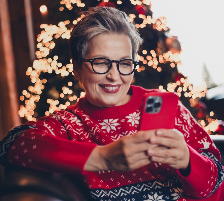 Eine grauhaarige Frau mittleren Alters mit Brille lächelt, während sie auf einem Sessel in rotem Weihnachtspullover ein Smartphone hält. | © AdobeStock/deagreez