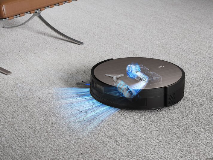 Ein runder Saugroboter mit visualisierter Technik saugt Haare und Staub von einem hellgrauen Teppich vor einem Ledersessel. | © Ecovacs