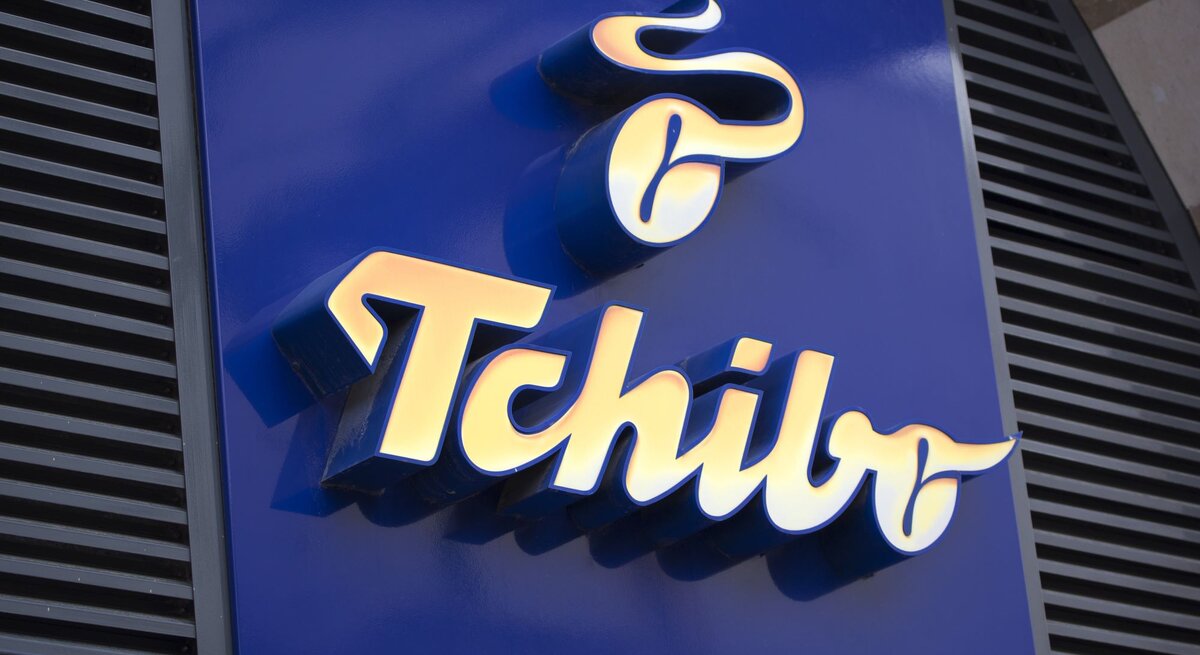 Golden leuchtendes Tchibo-Logo auf glänzend blauer Fassade, umgeben von schwarzen Lamellen und einem hellen Gebäuderand.