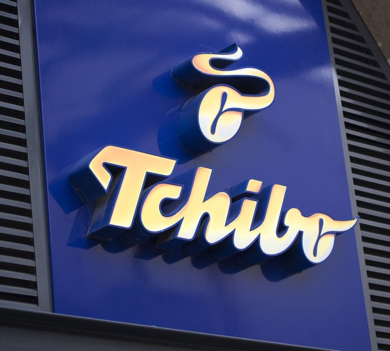 Golden leuchtendes Tchibo-Logo auf glänzend blauer Fassade, umgeben von schwarzen Lamellen und einem hellen Gebäuderand. | © AdobeStock/	asbe