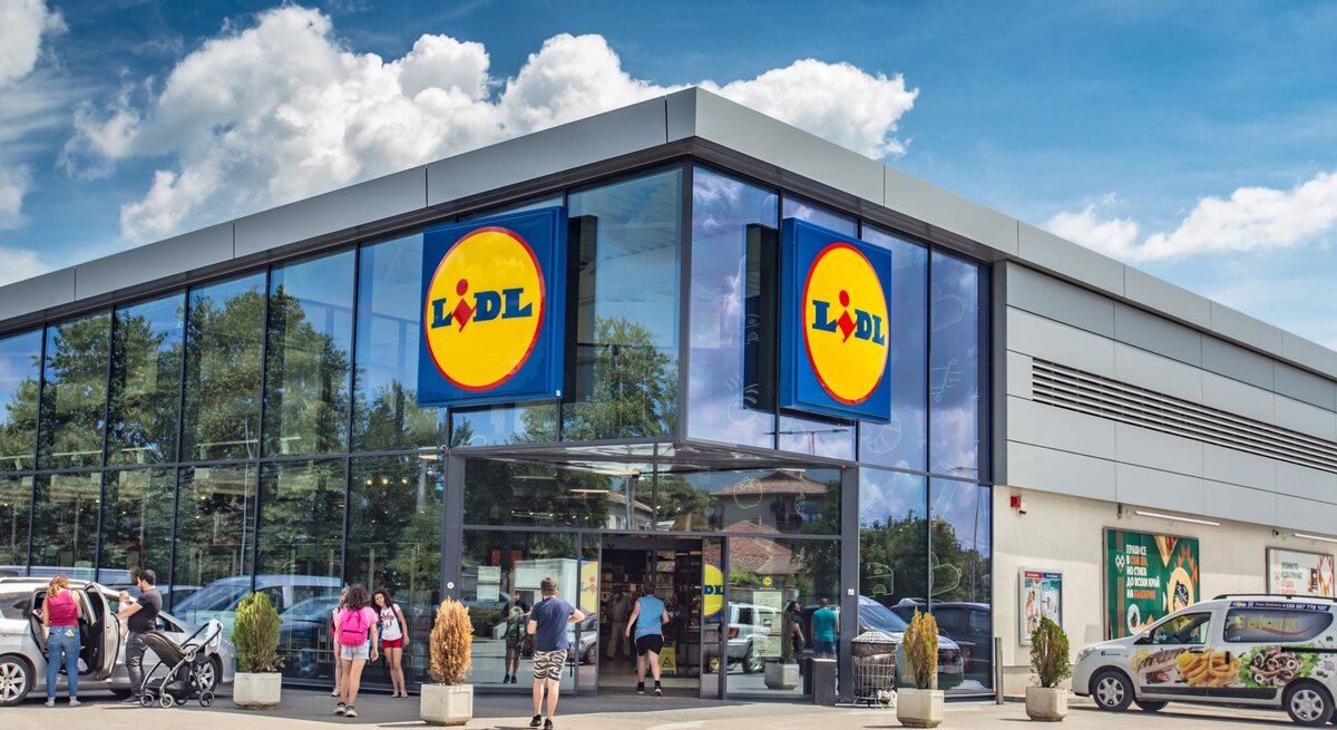 Das Foto zeigt den Eingangsbereich einer Lidl-Filiale an einem sonnigen Tag. Zwei große Schilder mit dem Lidl-Logo sind deutlich an der Fassade angebracht, was die Markenidentität des Discounters unterstreicht. Vor dem Geschäft sieht man einige Kunden, die das Geschäft betreten oder verlassen, was auf einen gewöhnlichen und belebten Einkaufstag hindeutet. Die geparkten Autos und das helle Wetter tragen zu der alltäglichen Szene eines Supermarktes bei, der für seine wirtschaftlichen Preise und ein vielfältiges Angebot bekannt ist.
