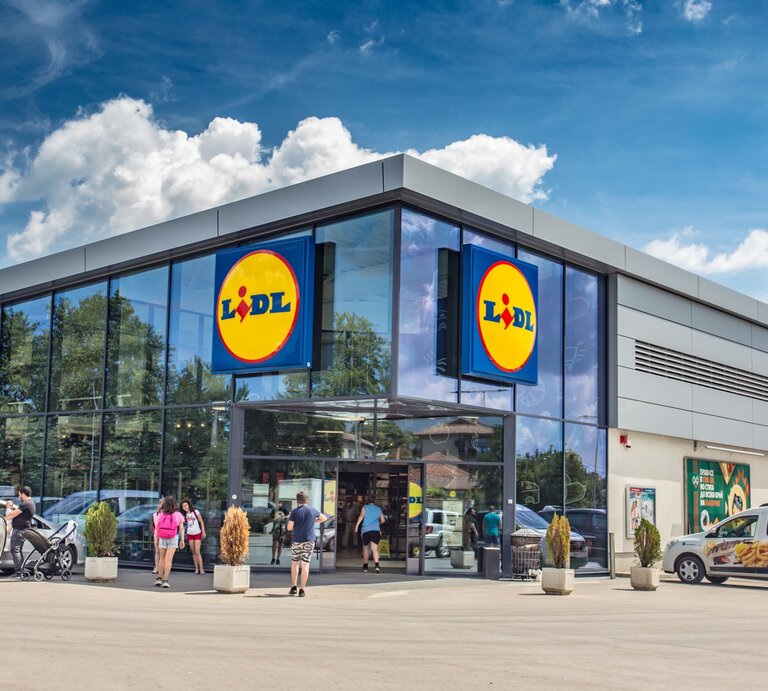 Das Foto zeigt den Eingangsbereich einer Lidl-Filiale an einem sonnigen Tag. Zwei große Schilder mit dem Lidl-Logo sind deutlich an der Fassade angebracht, was die Markenidentität des Discounters unterstreicht. Vor dem Geschäft sieht man einige Kunden, die das Geschäft betreten oder verlassen, was auf einen gewöhnlichen und belebten Einkaufstag hindeutet. Die geparkten Autos und das helle Wetter tragen zu der alltäglichen Szene eines Supermarktes bei, der für seine wirtschaftlichen Preise und ein vielfältiges Angebot bekannt ist. | © Adobe Stock/ Dinko