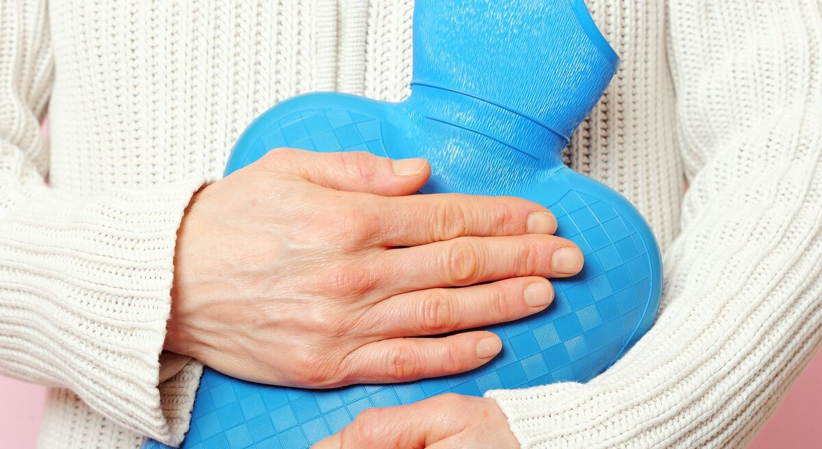 Eine Person hält eine blaue Wärmflasche vor ihrem Bauch, trägt einen weißen Strickpullover und hat blasse Hände.