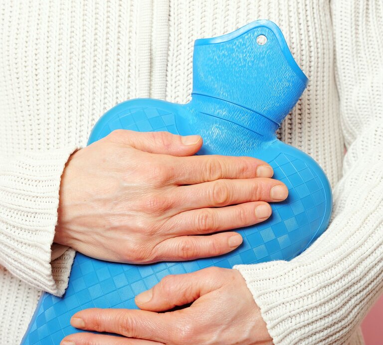 Eine Person hält eine blaue Wärmflasche vor ihrem Bauch, trägt einen weißen Strickpullover und hat blasse Hände. | © AdobeStock/granata68