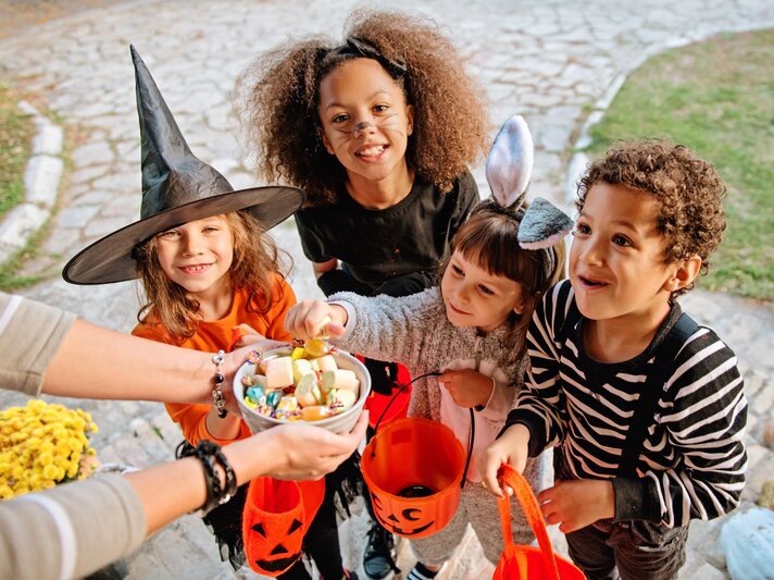 Vier fröhliche Kinder in Halloween-Kostümen halten orangefarbene Eimer und nehmen Süßigkeiten aus einer Schale entgegen. | © AdobeStock/Tijana