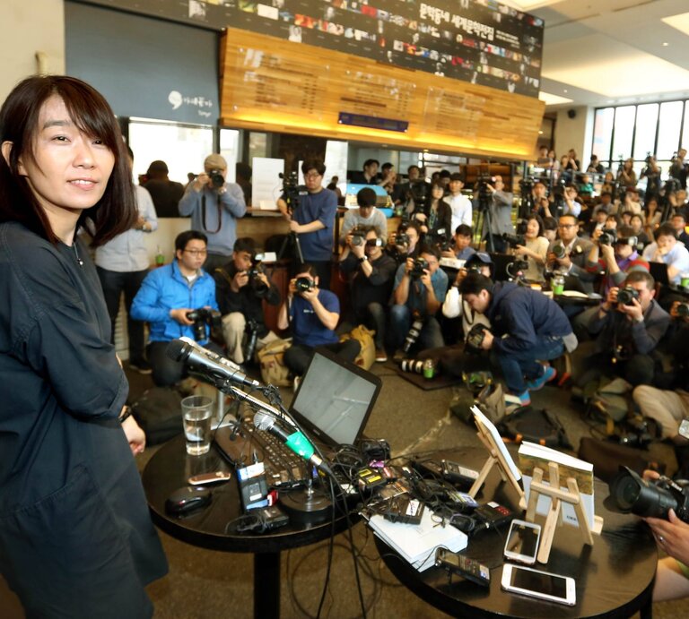 Eine Frau mittleren Alters mit schulterlangem braunem Haar steht lächelnd vor zahlreichen Fotografen in einem Konferenzraum. | © IMAGO/Newscom / Yonhap News