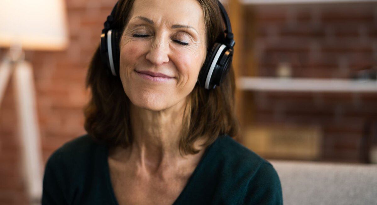 Eine lächelnde Frau mittleren Alters mit geschlossenen Augen genießt Musik über große Kopfhörer in einem gemütlichen Wohnzimmer.