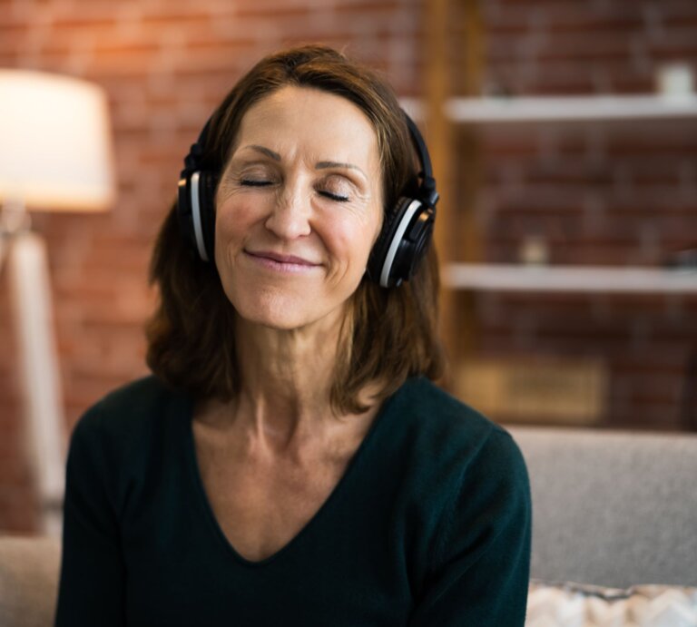 Eine lächelnde Frau mittleren Alters mit geschlossenen Augen genießt Musik über große Kopfhörer in einem gemütlichen Wohnzimmer. | © Adobe Stock/ Andrey Popov