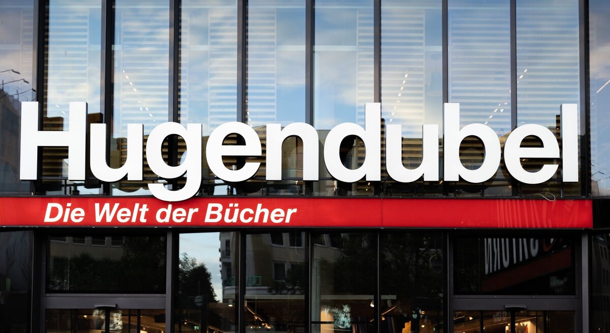 Große weiße Leuchtschrift "Hugendubel" mit rotem Slogan "Die Welt der Bücher" über der Glasfassade einer Buchhandlung.