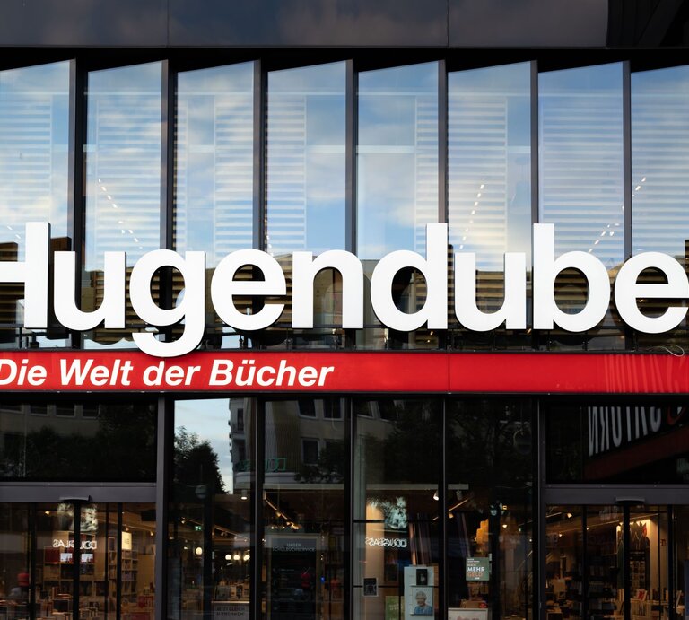 Große weiße Leuchtschrift "Hugendubel" mit rotem Slogan "Die Welt der Bücher" über der Glasfassade einer Buchhandlung. | © Adobe Stock/ 1take1shot