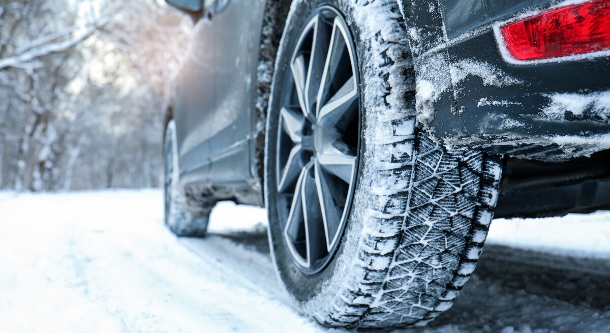 Ein dunkles Auto mit Schneereifen fährt auf einer verschneiten Straße durch einen winterlichen Wald mit kahlen Bäumen.