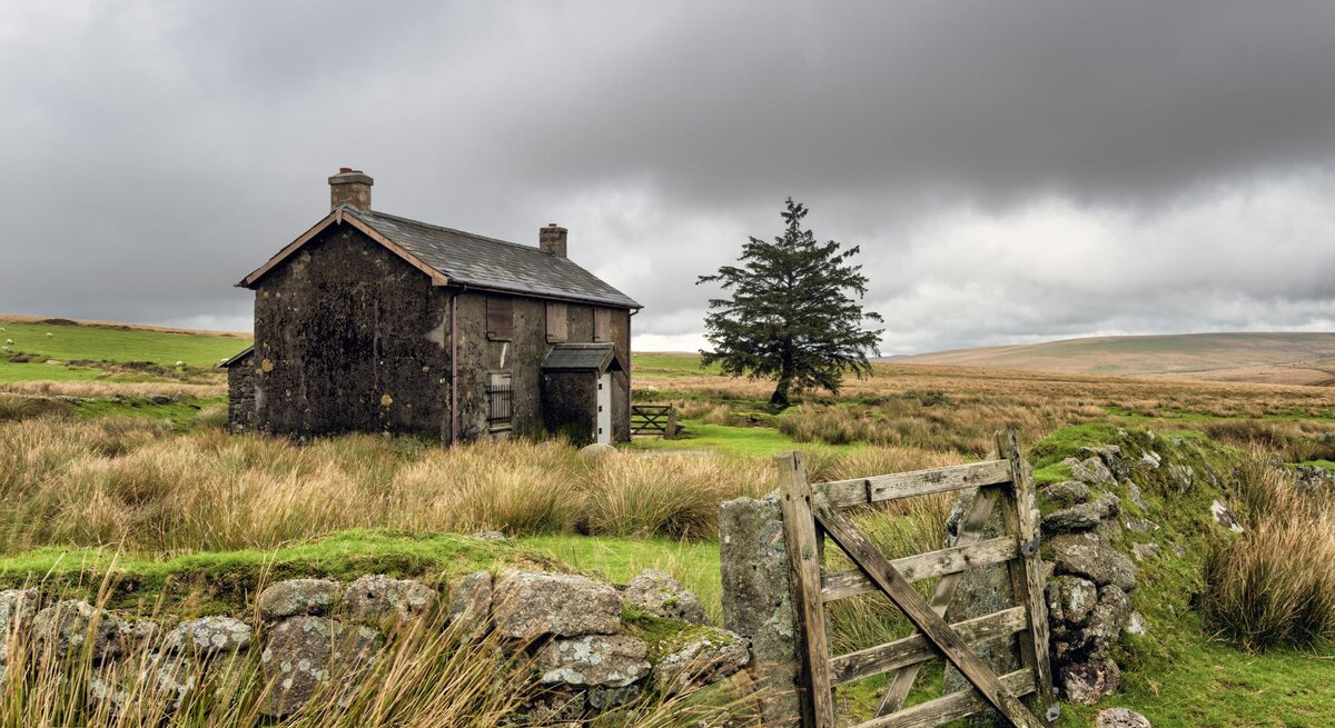 Ein verlassenes Steinhaus steht unter düsteren Wolken in einer abgelegenen, windgepeitschten Moorlandschaft neben einem Holzzaun.