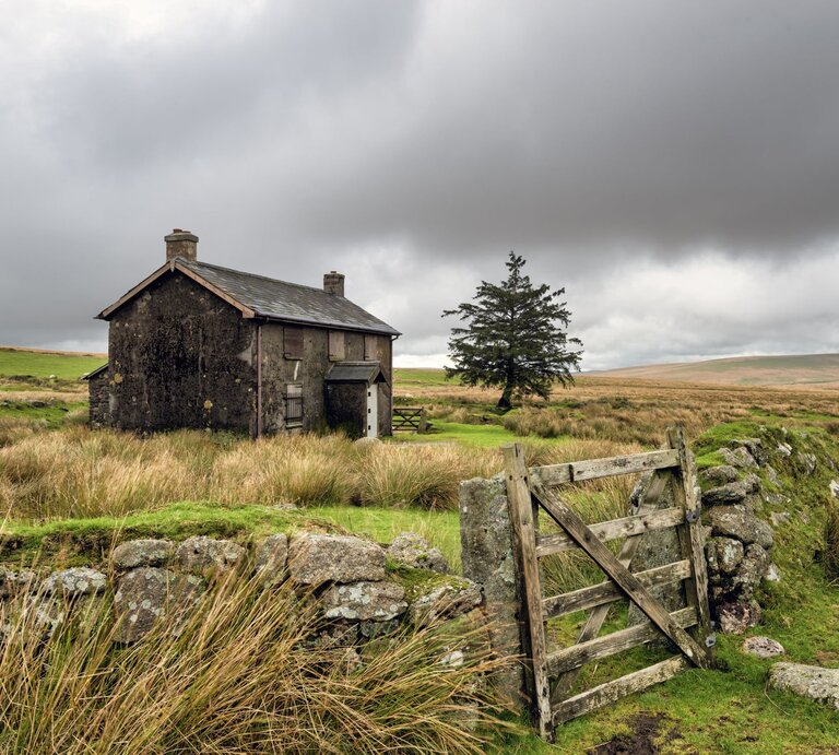 Ein verlassenes Steinhaus steht unter düsteren Wolken in einer abgelegenen, windgepeitschten Moorlandschaft neben einem Holzzaun. | © Shutterstock/Helen Hotson
