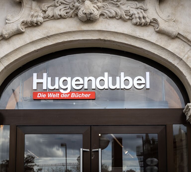 Eingang eines Hugendubel-Buchladens mit Schriftzug 'Die Welt der Bücher' unter einem steinernen Torbogen. | © Adobe Stock/ Dennis