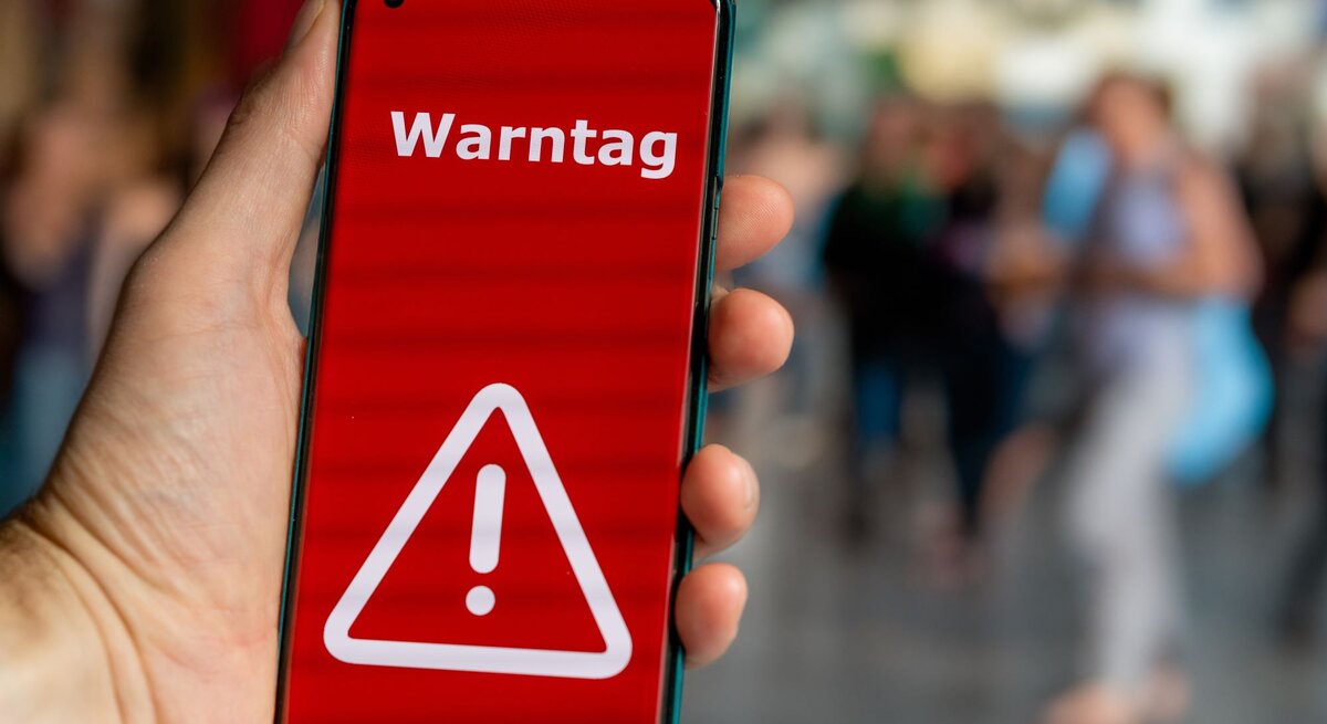 Hand hält Smartphone mit rotem Bildschirm und weißem Warnsymbol, darauf steht „Warntag“, im unscharfen Hintergrund Menschen.