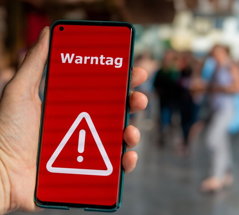 Hand hält Smartphone mit rotem Bildschirm und weißem Warnsymbol, darauf steht „Warntag“, im unscharfen Hintergrund Menschen. | © Adobe Stock/ Fokussiert