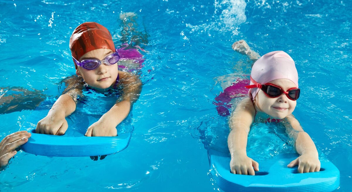 Zwei Kinder mit badekappe und schwimmbrille werden mit Schwimmbrettern an das Wasser gewöhnt.