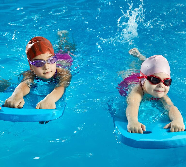 Zwei Kinder mit badekappe und schwimmbrille werden mit Schwimmbrettern an das Wasser gewöhnt. | © gettyimages/	Mariakray