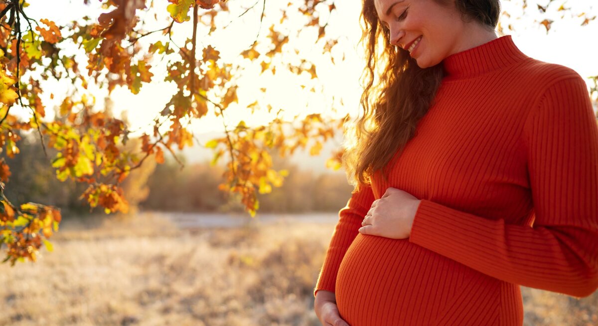 Junge schwangere Frau in einem orangefarbenen Kleid lächelt glücklich und hält ihren Bauch in einer herbstlichen Landschaft.