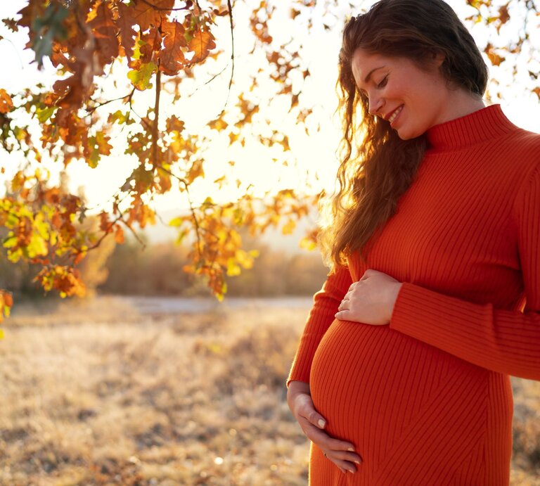 Junge schwangere Frau in einem orangefarbenen Kleid lächelt glücklich und hält ihren Bauch in einer herbstlichen Landschaft. | © Getty Images/SrdjanPav