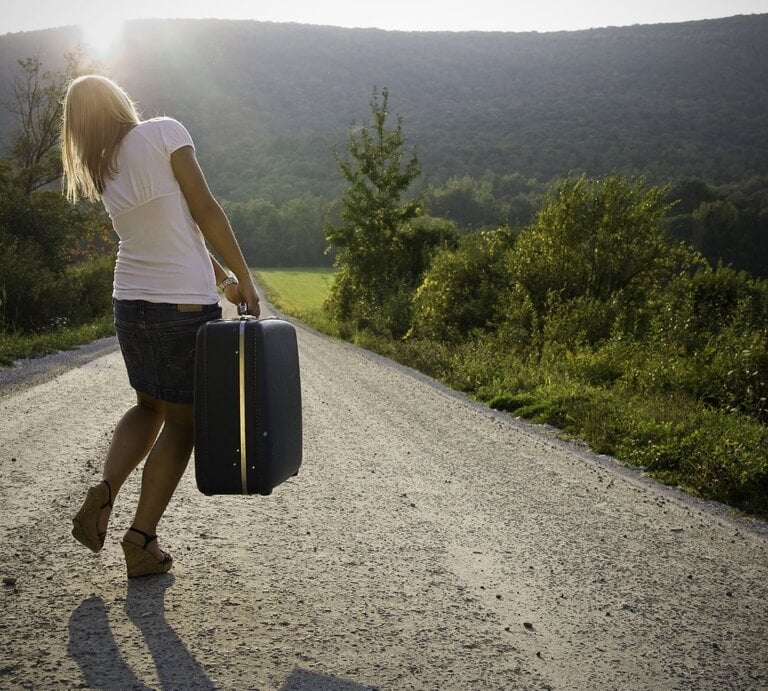 Eine blonde Frau in Jeansrock und Sandalen trägt einen Koffer auf einer sonnigen Schotterstraße inmitten einer grünen Hügellandschaft. | © Getty Images/cglade