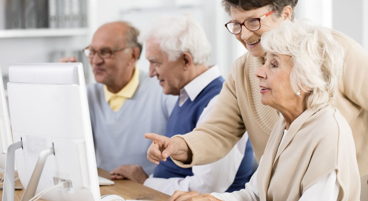 Eine ältere Frau zeigt einer anderen Frau etwas auf einem Computerbildschirm. Sie freuen sich. Zwei Männer im Hintergrund.