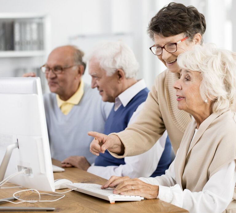 Eine ältere Frau zeigt einer anderen Frau etwas auf einem Computerbildschirm. Sie freuen sich. Zwei Männer im Hintergrund. | © gettyimages/	KatarzynaBialasiewicz