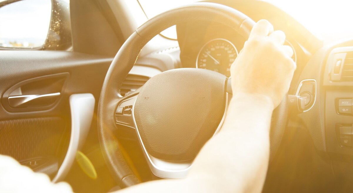 Eine Hand am Lenkrad eines Autos, in das warme, gleißende Sonnenlicht strahlt.