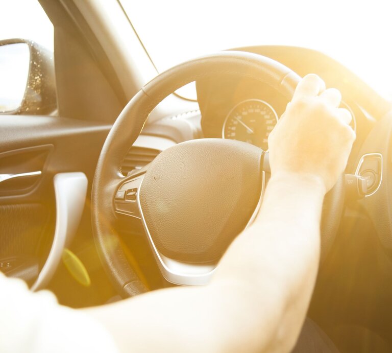 Eine Hand am Lenkrad eines Autos, in das warme, gleißende Sonnenlicht strahlt. | © Getty Images/eternalcreative