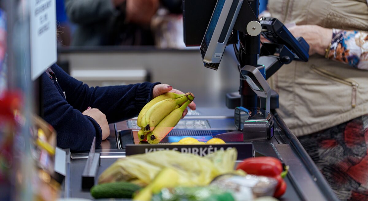 Gelbe Bananen werden an einer belebten Supermarktkasse gescannt, vorne liegen Gurke und Paprika, hinten Kleidung und Hände sichtbar.