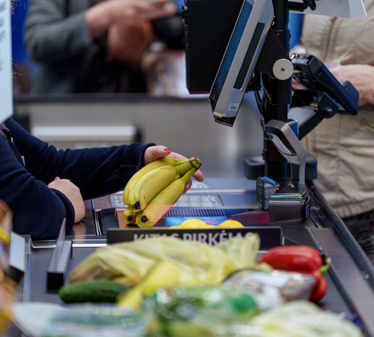 Gelbe Bananen werden an einer belebten Supermarktkasse gescannt, vorne liegen Gurke und Paprika, hinten Kleidung und Hände sichtbar. | © Adobe Stock/ Rokas