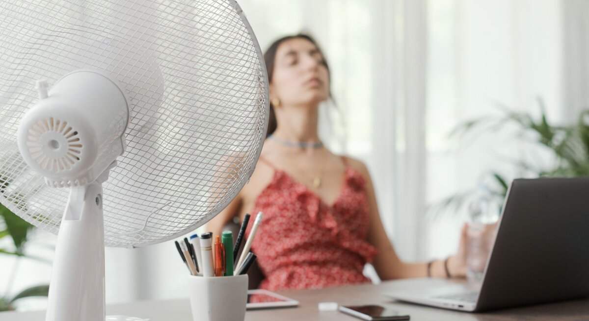Junge Frau in rotem Sommerkleid sitzt erschöpft vor einem Laptop, während ein Ventilator auf sie gerichtet ist.