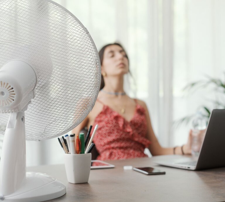Junge Frau in rotem Sommerkleid sitzt erschöpft vor einem Laptop, während ein Ventilator auf sie gerichtet ist. | © Adobe Stock/StockPhotoPro