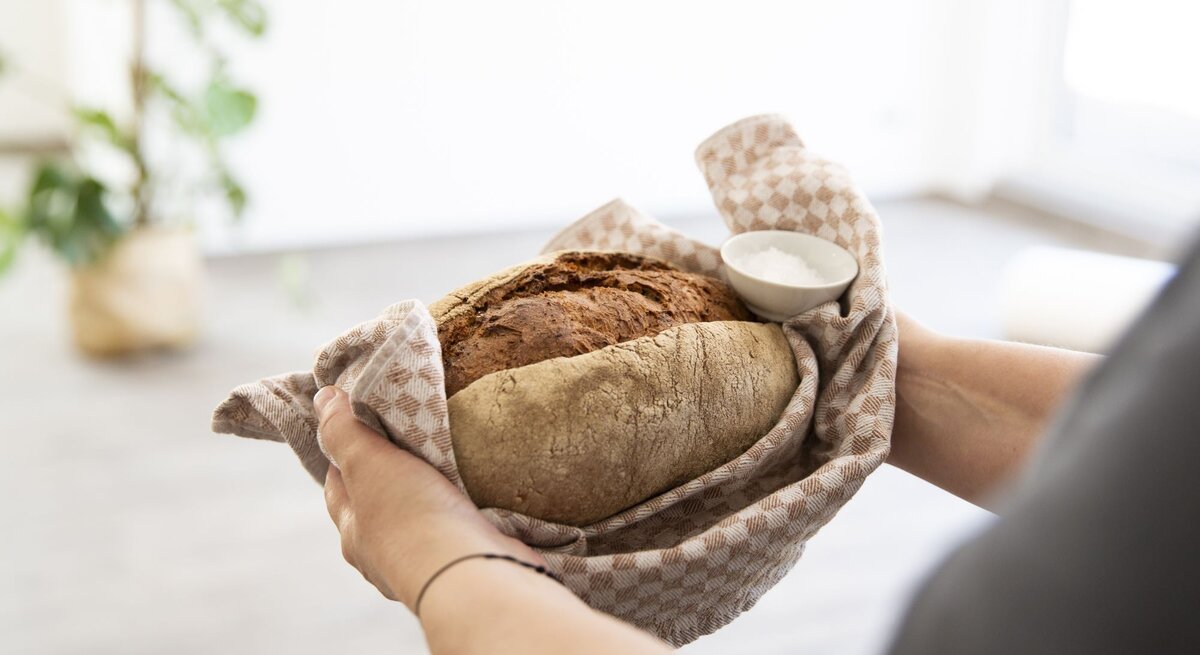 Zwei Hände halten ein frisch gebackenes Brot in einem beige karierten Tuch, daneben eine Schale mit Salz, vor unscharfem Hintergrund.