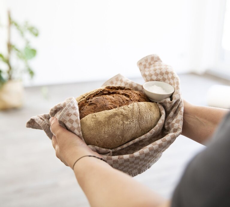 Zwei Hände halten ein frisch gebackenes Brot in einem beige karierten Tuch, daneben eine Schale mit Salz, vor unscharfem Hintergrund. | © Getty Images/Westend61
