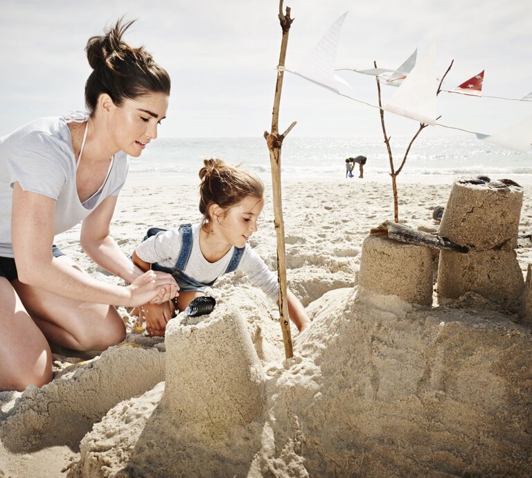 Eine junge Frau mit braunem Haar und ein Mädchen in Latzhose bauen konzentriert eine Sandburg am sonnigen Strand. | © Getty Images/Uwe Krejci