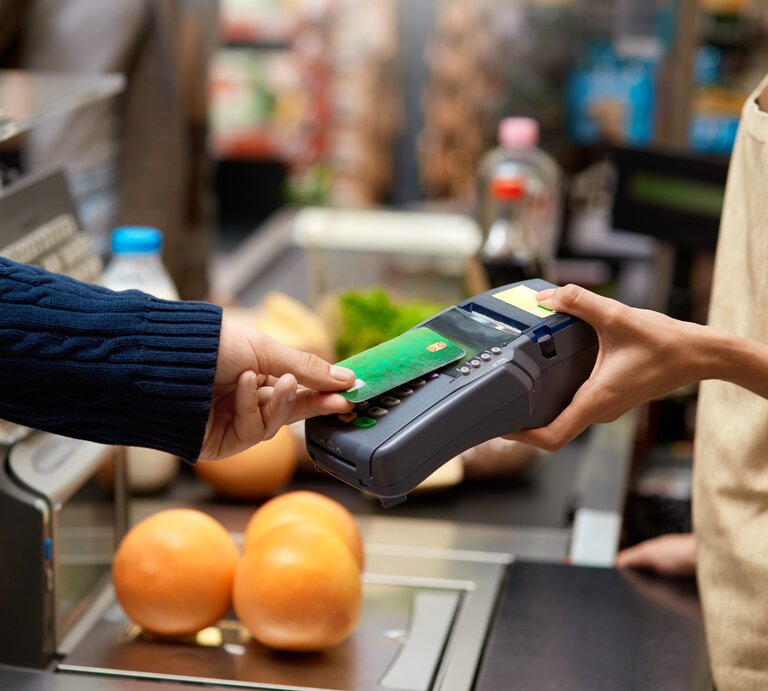 Eine Person in blauem Pullover hält eine grüne Kreditkarte über ein Kartenterminal, im Hintergrund sind frische Lebensmittel zu sehen. | © Adobe Stock/ Viktoriia