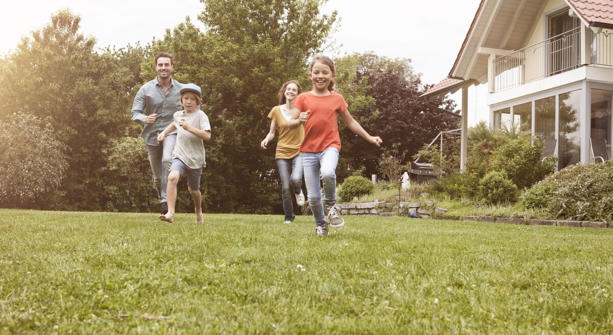 Eine Familie mit zwei Kindern rennt lachend über eine grüne Wiese vor einem modernen Haus an einem sonnigen Tag.