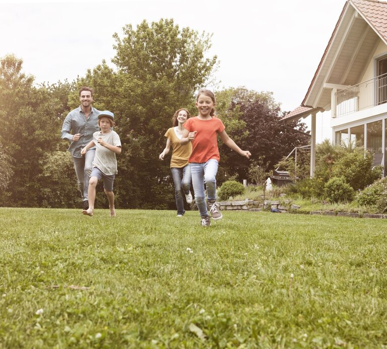 Eine Familie mit zwei Kindern rennt lachend über eine grüne Wiese vor einem modernen Haus an einem sonnigen Tag. | © Getty Images/Westend61