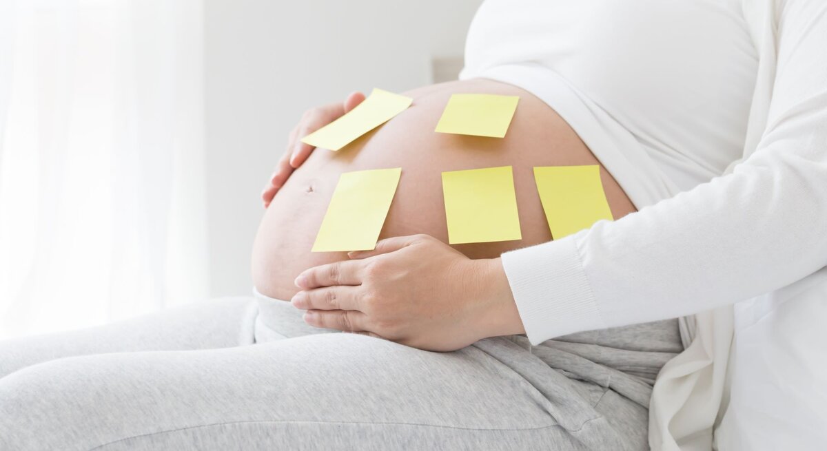 Schwangere Frau in grauer Hose und weißem Oberteil sitzt entspannt und hat gelbe Notizzettel auf ihrem Bauch.