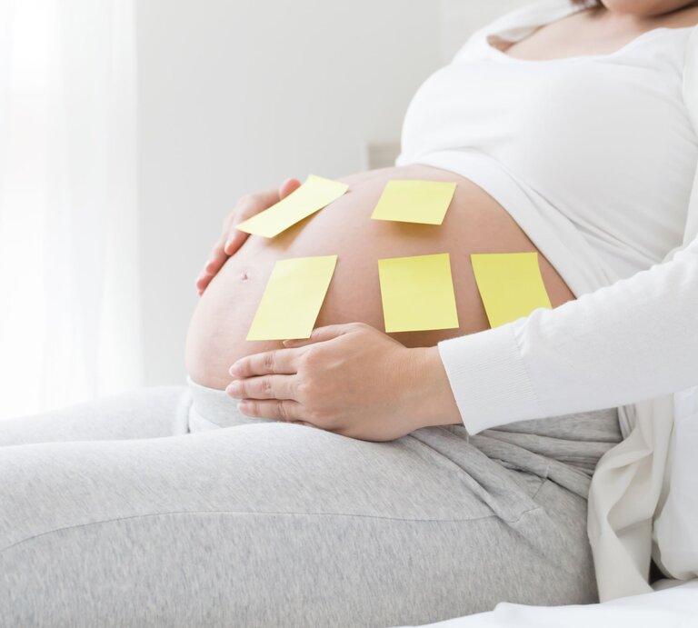 Schwangere Frau in grauer Hose und weißem Oberteil sitzt entspannt und hat gelbe Notizzettel auf ihrem Bauch. | © Getty Images/comzeal