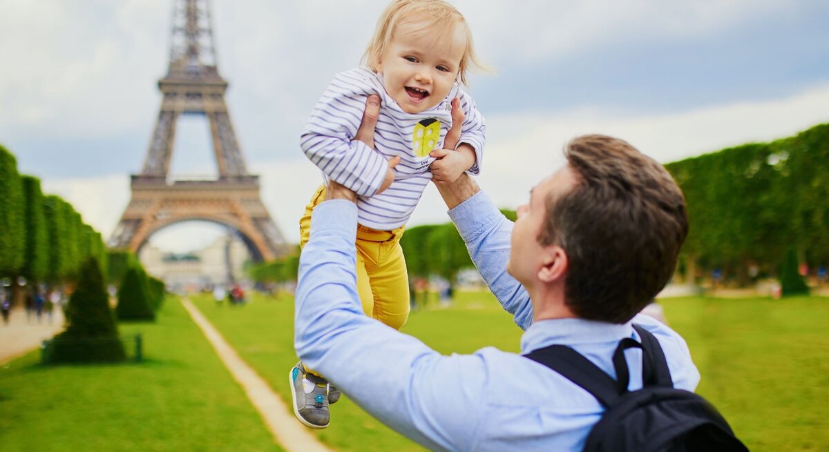 Ein lachender Vater hebt sein fröhliches Kleinkind vor dem Eiffelturm in Paris in die Luft, auf einer grünen Wiese.