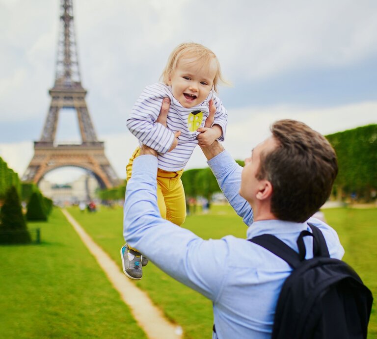Ein lachender Vater hebt sein fröhliches Kleinkind vor dem Eiffelturm in Paris in die Luft, auf einer grünen Wiese. | © Adobe Stock/Ekaterina Pokrovsky