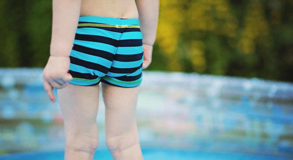Ein Kleinkind in blau-schwarz gestreifter Badehose steht am Rand eines Planschbeckens mit verschwommenem grünen Hintergrund.