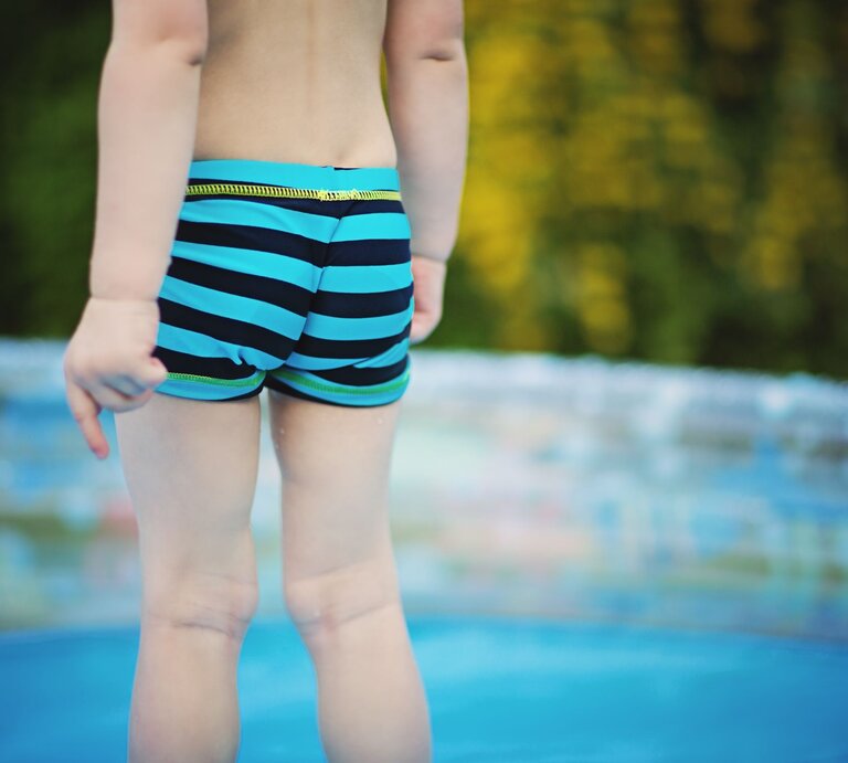 Ein Kleinkind in blau-schwarz gestreifter Badehose steht am Rand eines Planschbeckens mit verschwommenem grünen Hintergrund. | © Getty Images/Tatjana Kaufmann