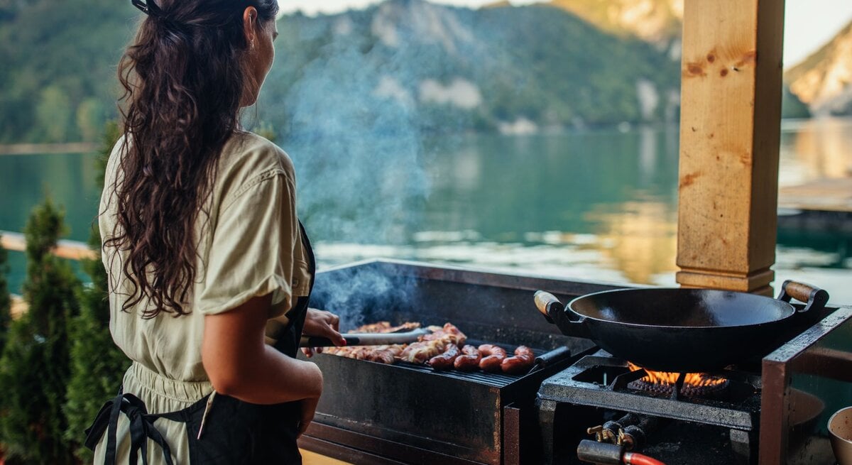 Eine junge Frau mit langen braunen Haaren grillt am Abend Würstchen an einem See mit Bergkulisse im Hintergrund.