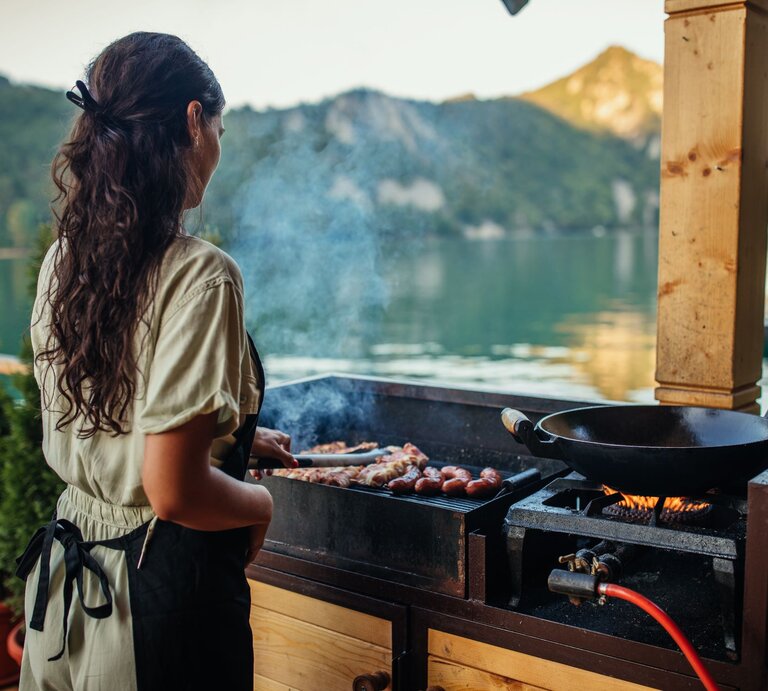 Eine junge Frau mit langen braunen Haaren grillt am Abend Würstchen an einem See mit Bergkulisse im Hintergrund. | © Shutterstock/bbernard