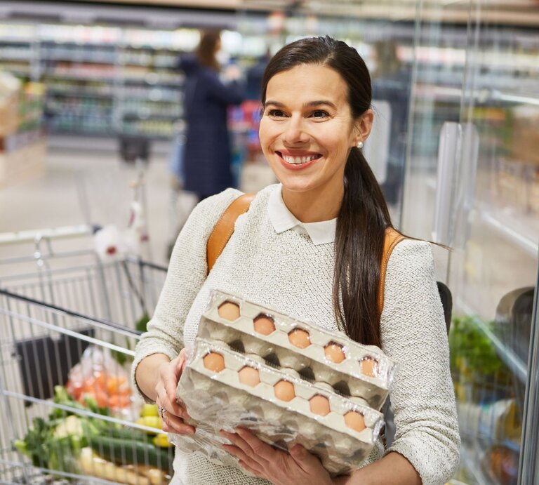 Eine junge Frau mit braunem Haar und weißem Pullover hält lächelnd zwei Eierkartons in einem Discounter. | © Adobe Stock/Robert Kneschke