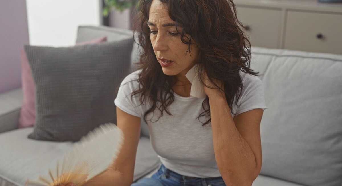 Eine Frau mit dunklem, lockigem Haar trägt Jeans und weißes T-Shirt, fächelt sich sitzend in einem modernen Wohnzimmer Luft zu.