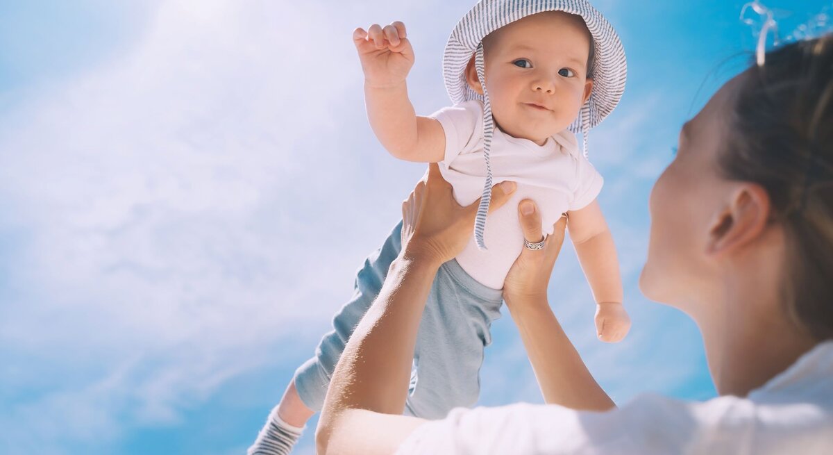 Junge Frau hebt lächelnd ein Baby mit gestreiftem Sonnenhut und weißem T-Shirt in den strahlend blauen Himmel.